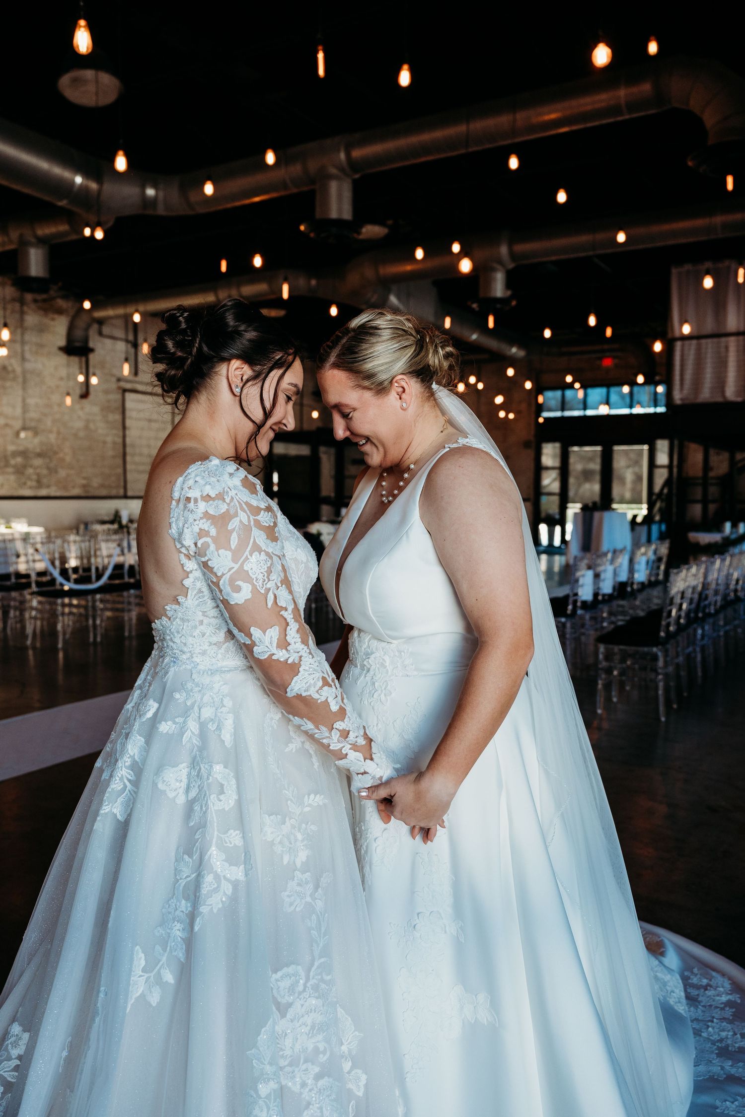 Two brides in elegant white gowns stand close together under romantic venue lighting.
