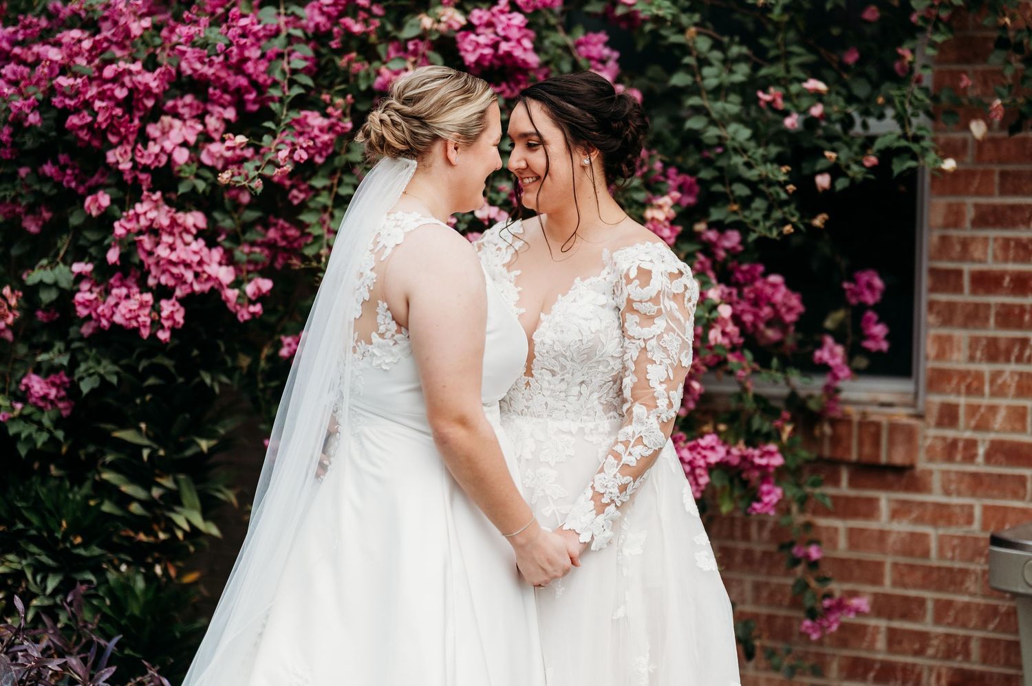 A couple in wedding attire embraces near a brick wall with vibrant pink flowers in the background.