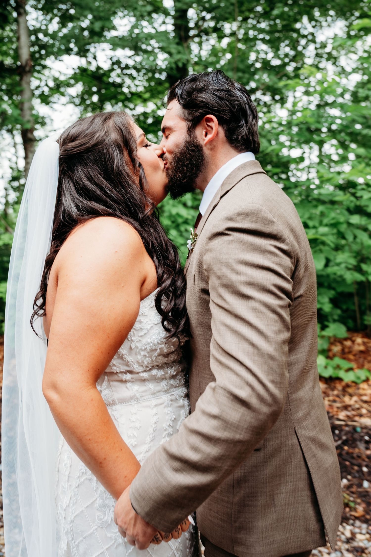 A bride and groom share a romantic kiss outdoors, holding hands, with lush green trees in the background.