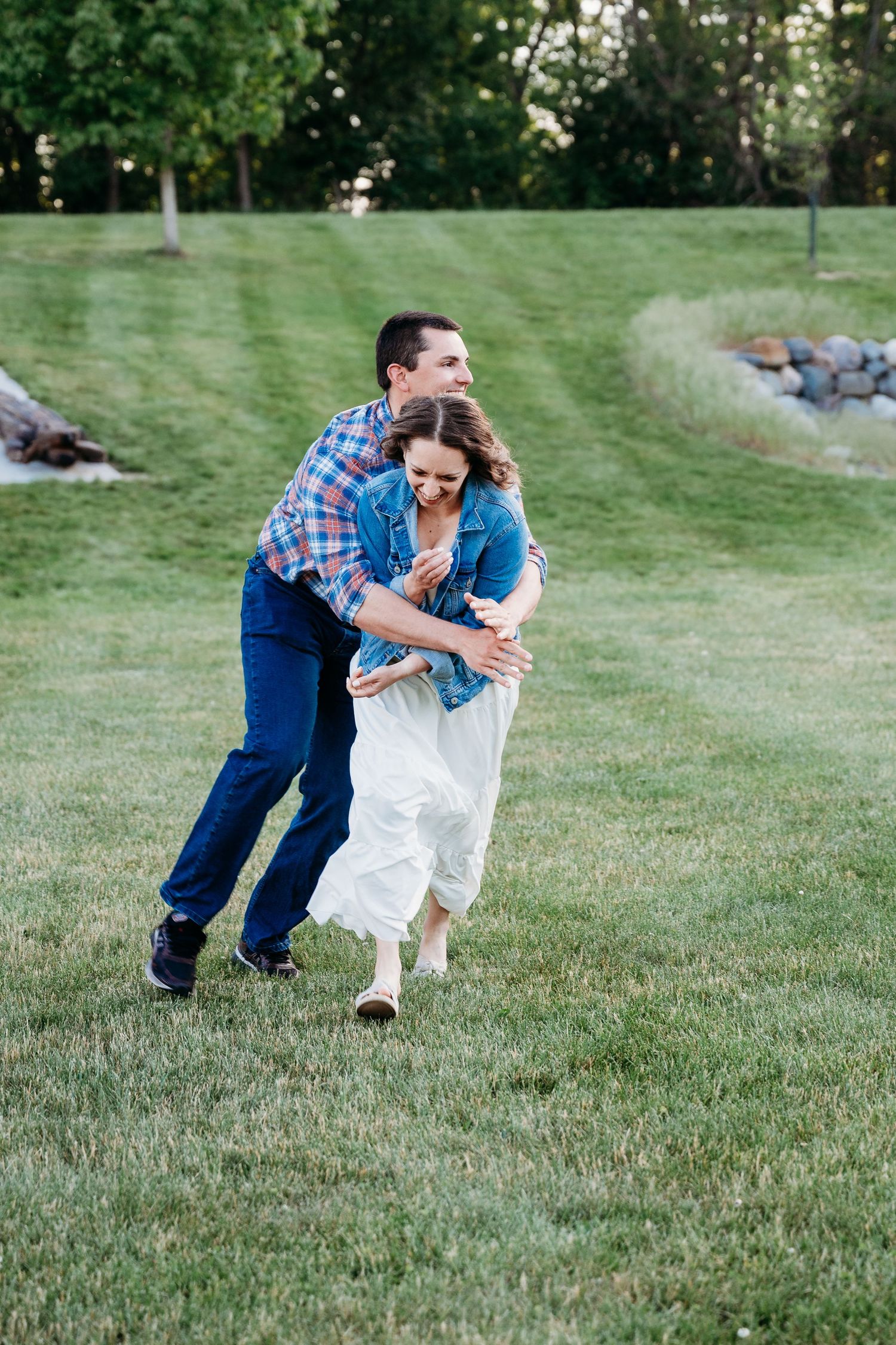 A joyful couple plays in a lush green park, man in plaid shirt hugging woman in white dress.