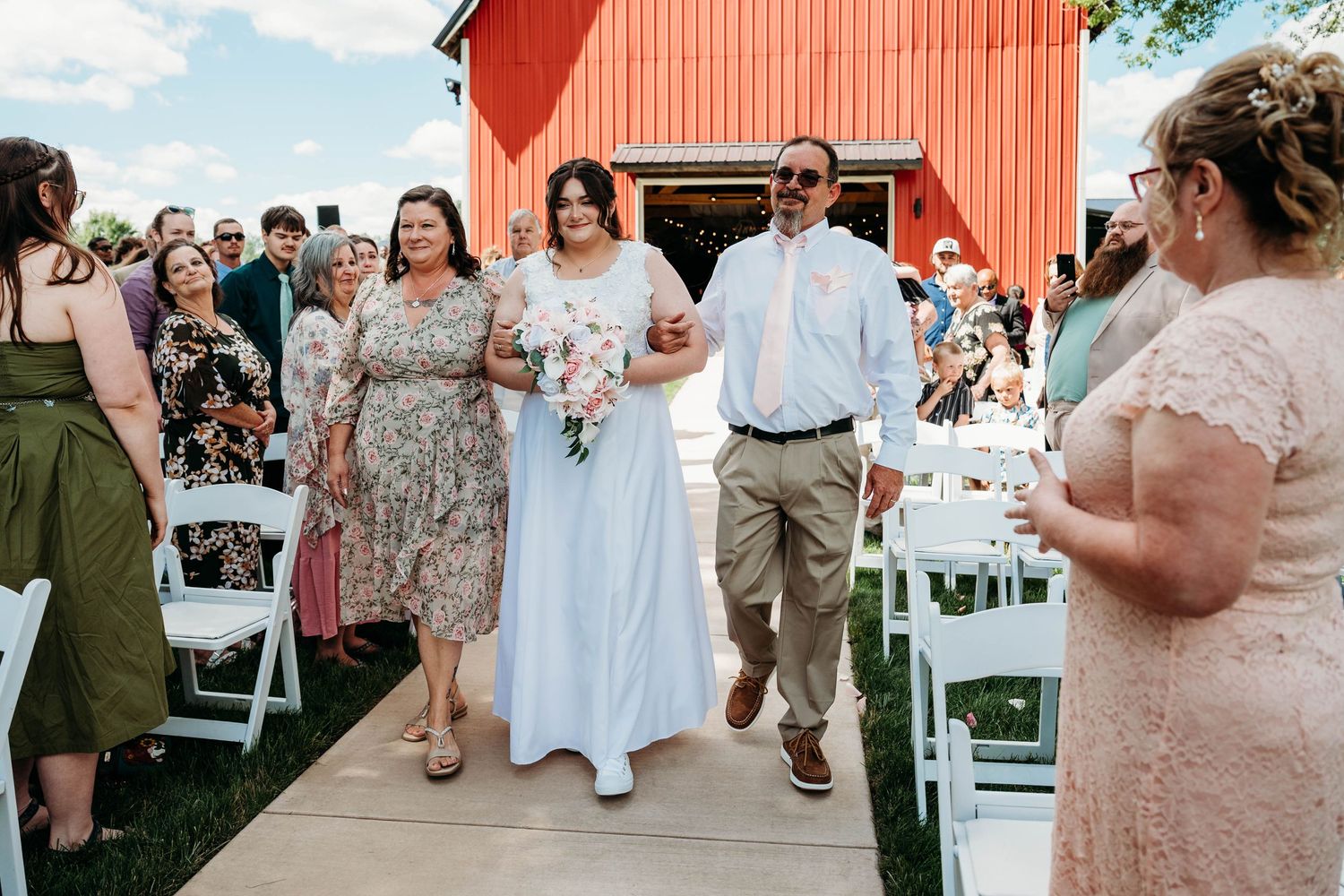 Bride walks down the aisle with parents at outdoor barn wedding ceremony with red barn backdrop.