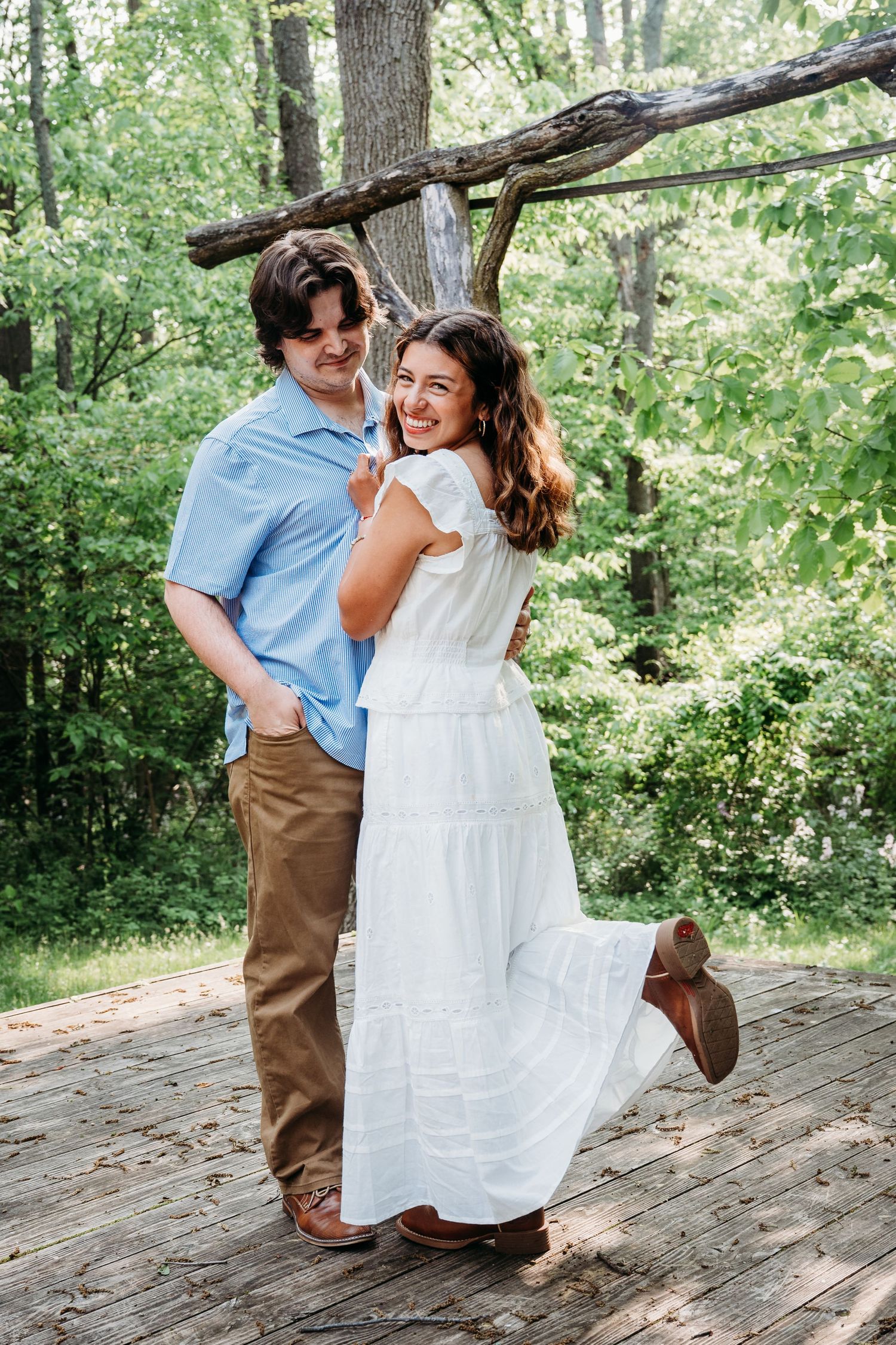 A joyful couple laughs together on a wooden deck in a lush forest setting, she wearing a flowing white dress.