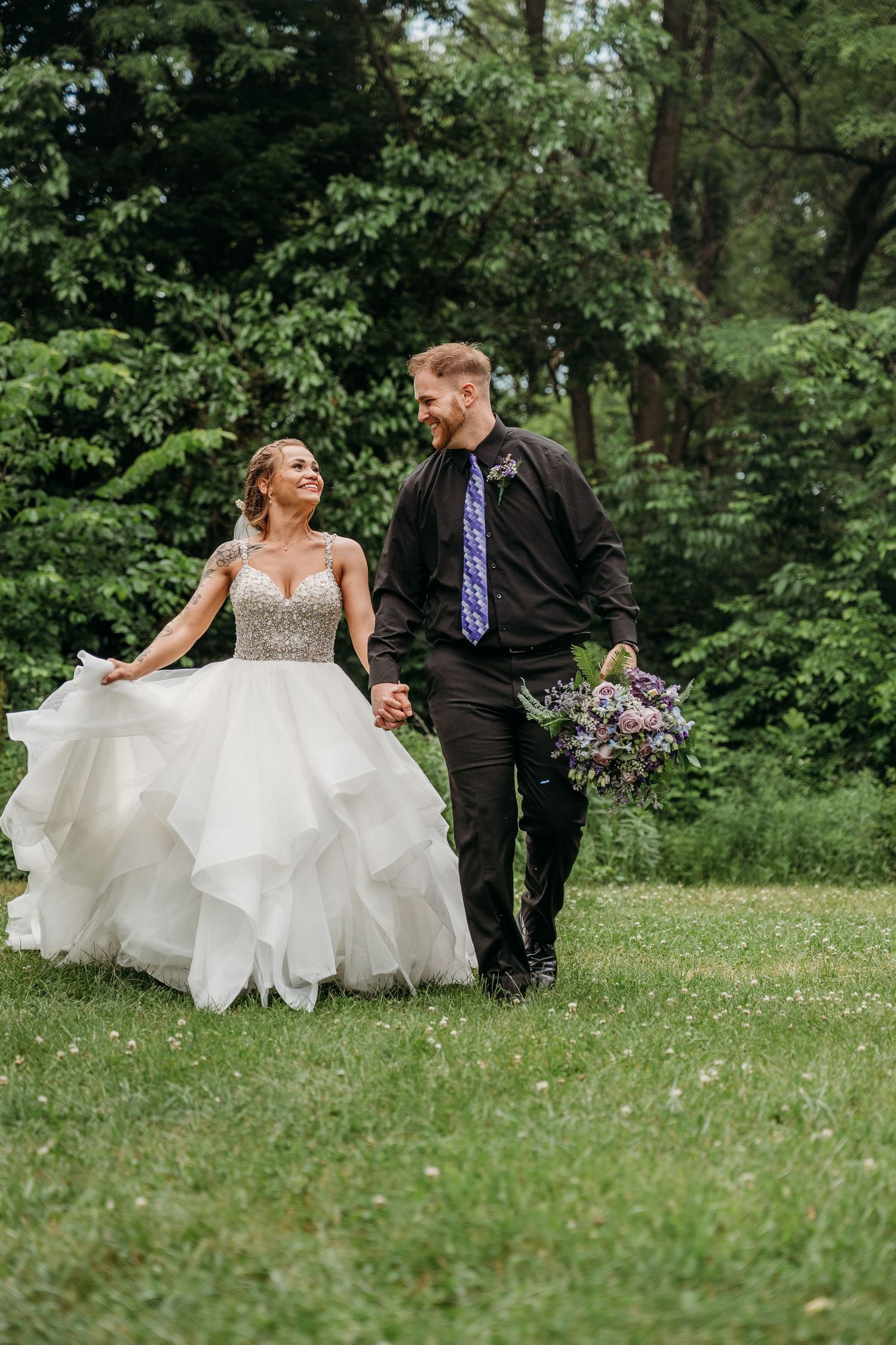 A bride and groom walk together across a lush green lawn surrounded by trees on their wedding day.