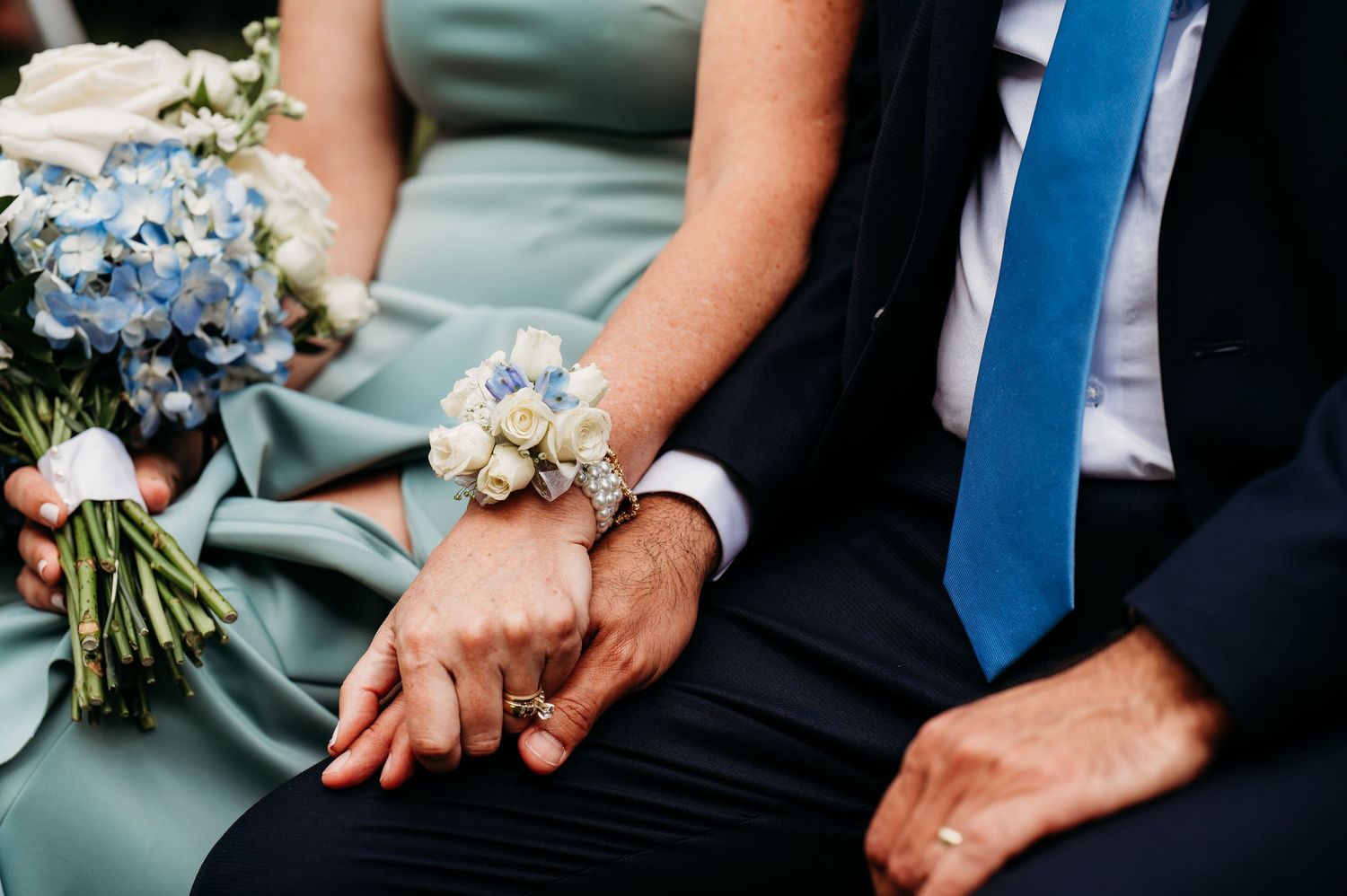 Close-up of hands holding delicate blue and white floral bouquets at a wedding ceremony against sage green attire.