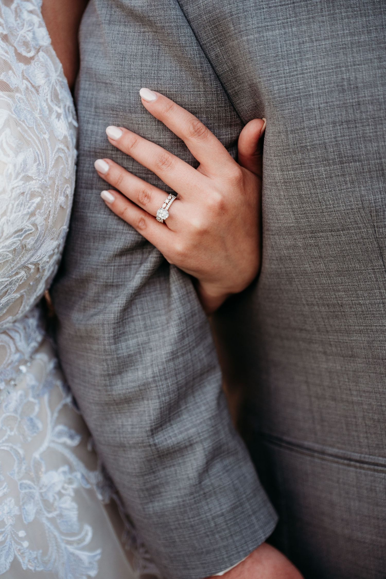 Close-up of a hand wearing an engagement ring resting on a gray suit jacket next to white lace fabric.
