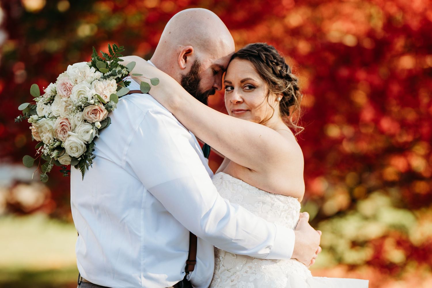 A romantic wedding couple embraces in front of vibrant autumn foliage while holding a white and peach floral bouquet.