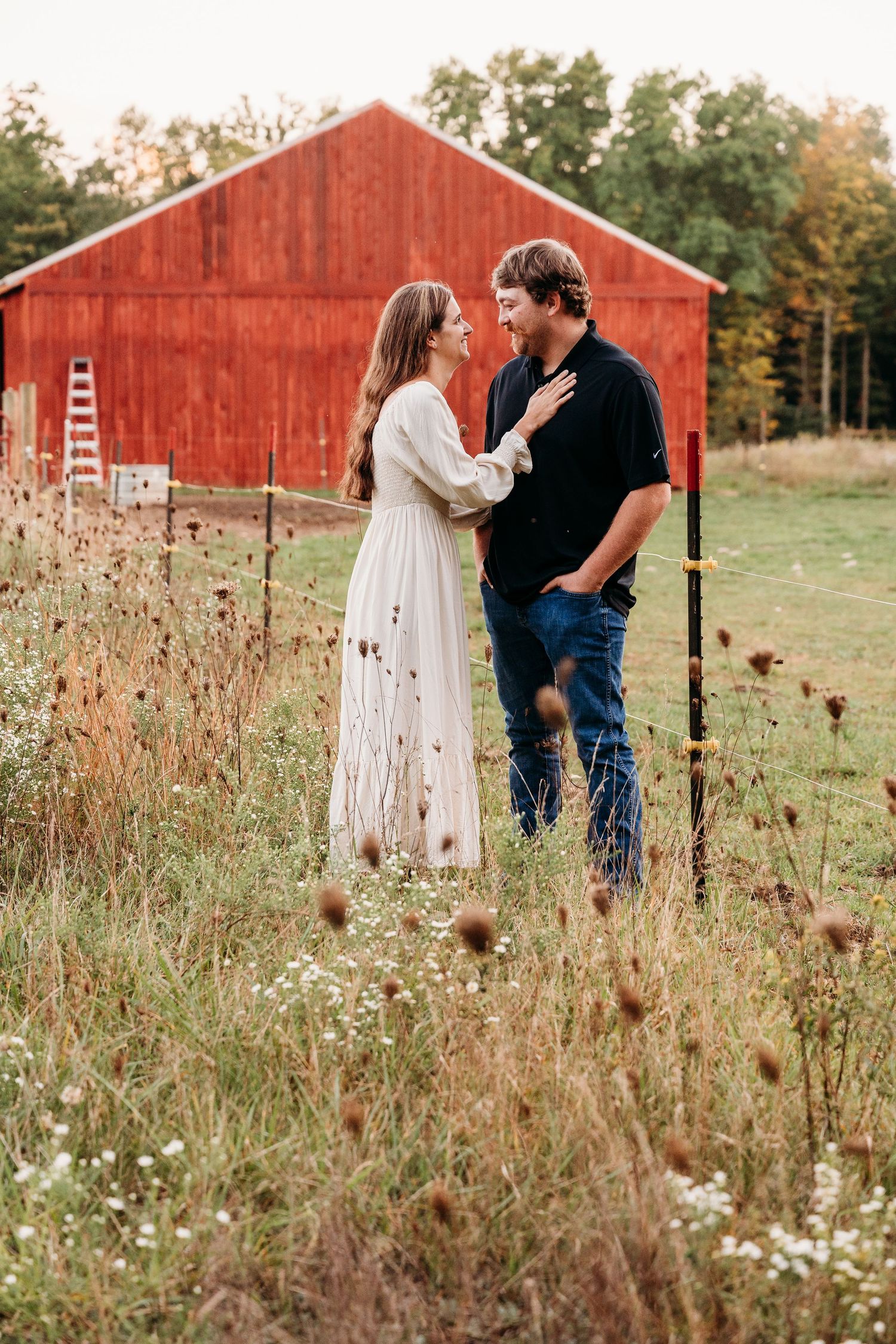 A couple shares a romantic moment in a rustic field with wildflowers and a red barn in the background.