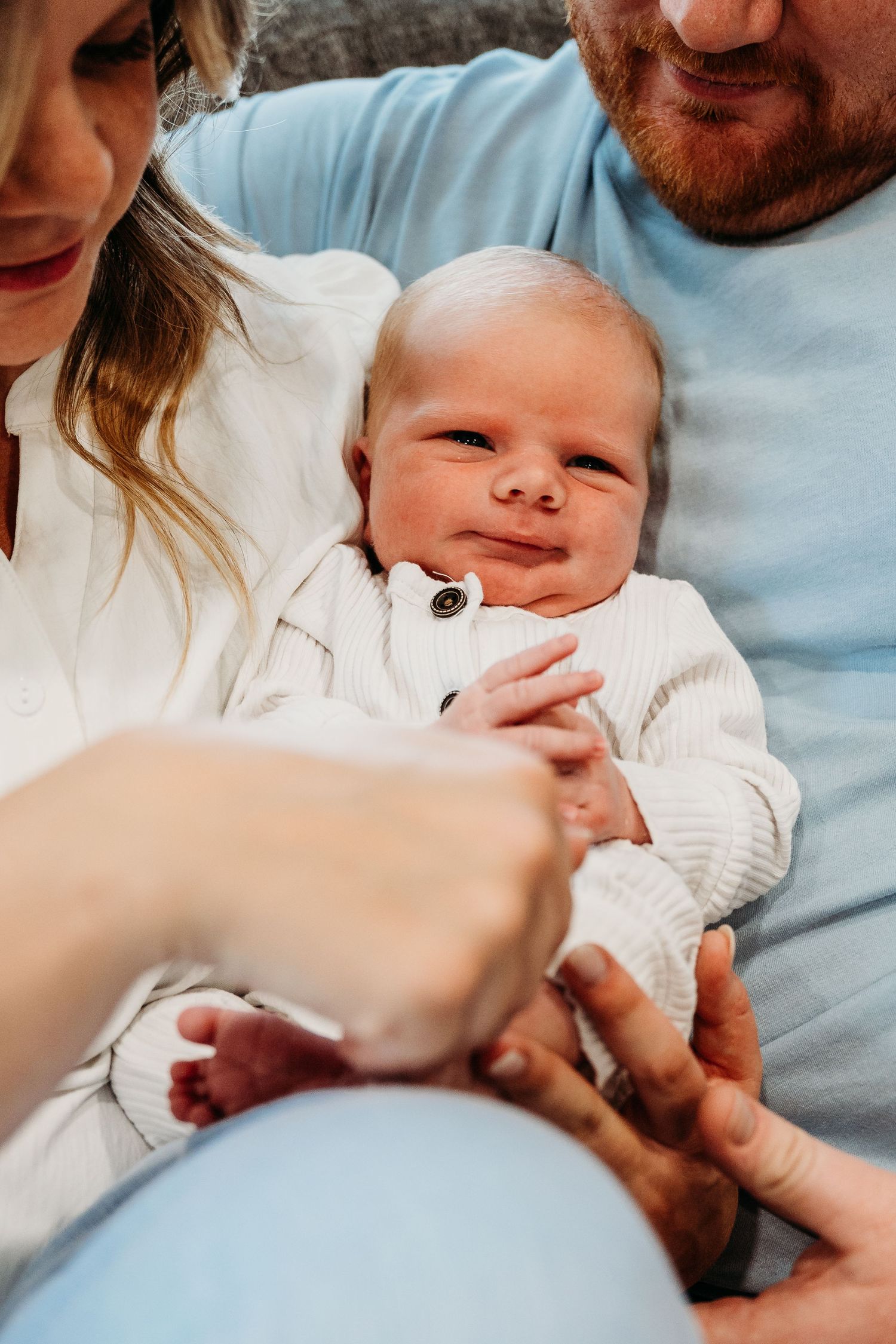 A baby in a white sweater smiles while being held by parents in a casual home setting.