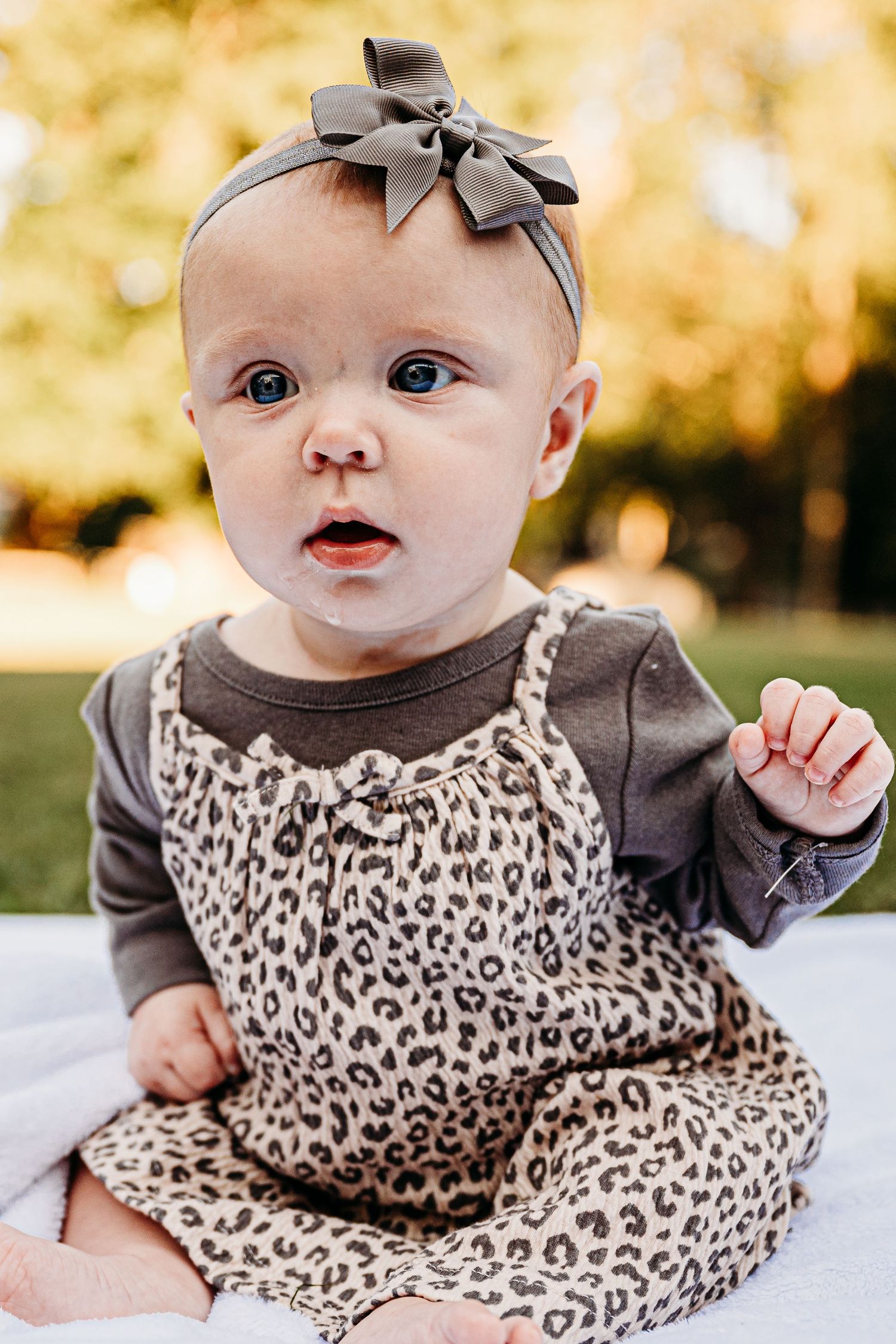 Baby wearing a leopard print dress and gray bow headband lying on grass during autumn.