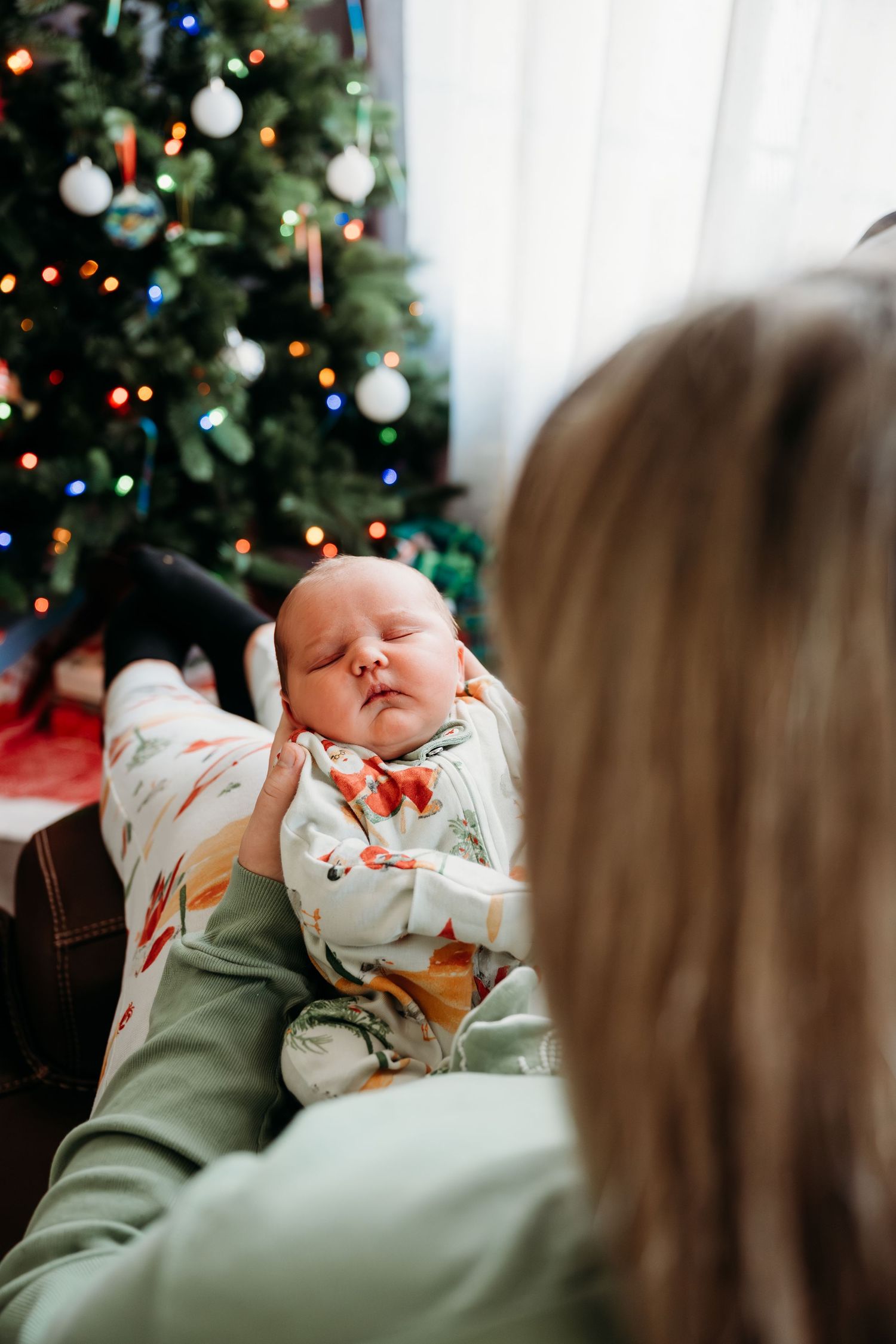 Baby in white onesie being held against green sweater with Christmas tree and ornaments in background.