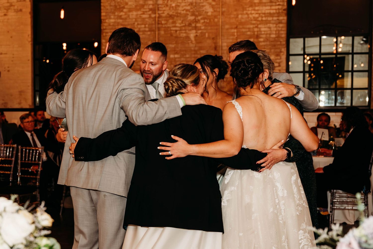 Wedding guests and couple share an intimate group hug during reception celebration in brick-walled venue.