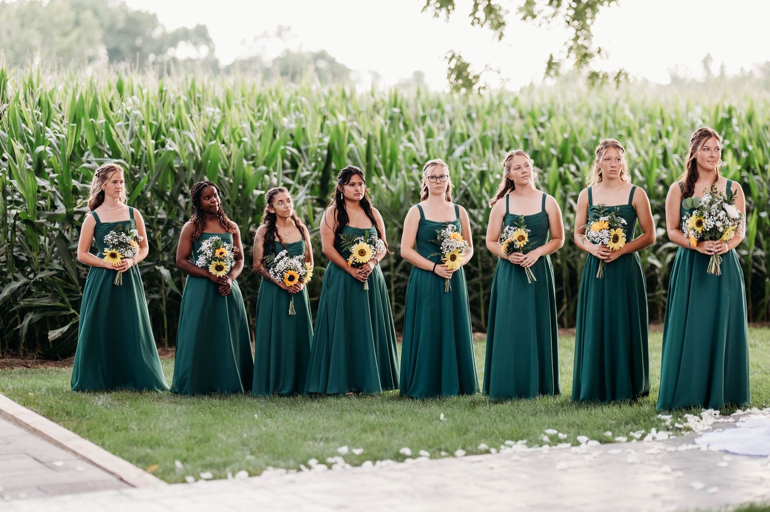 Seven bridesmaids in emerald green gowns holding sunflower bouquets pose outdoors near a lush cornfield.