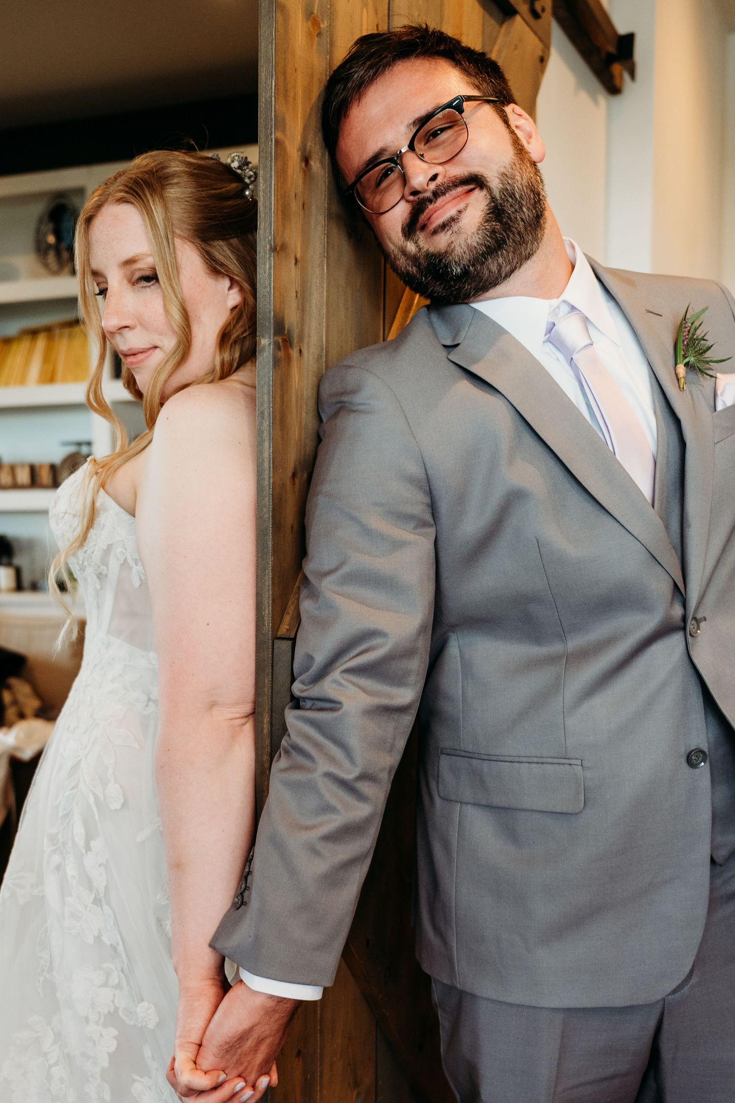 A bride and groom share a sweet moment while standing back to back against a wooden wall in their wedding attire.