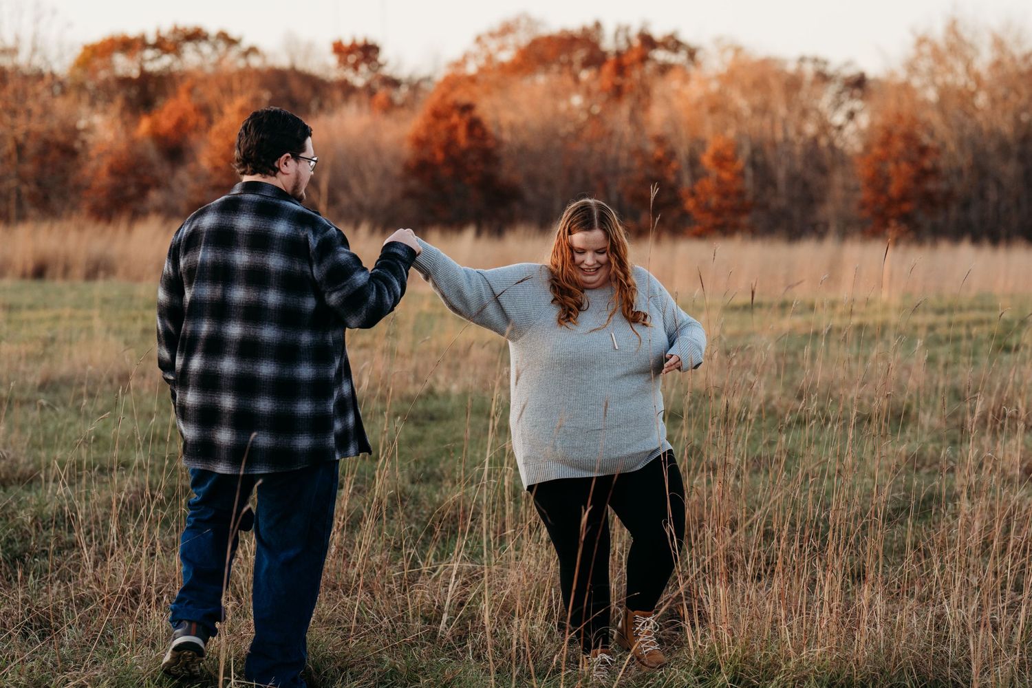 Two people share a playful moment in an autumn field with warm sunset light and tall grasses in the background.