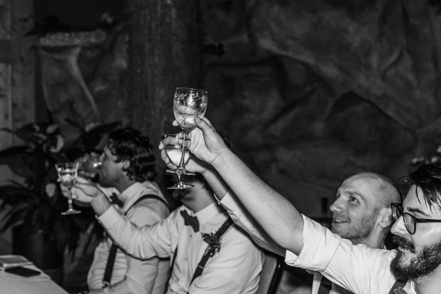 Black and white photo of guests raising wine glasses in a celebratory toast at a wedding reception.