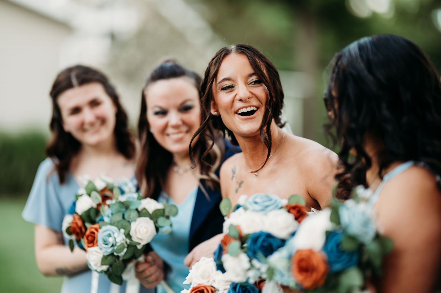 Bridesmaids in light blue dresses laughing and holding floral bouquets with white, orange, and blue flowers outdoors.