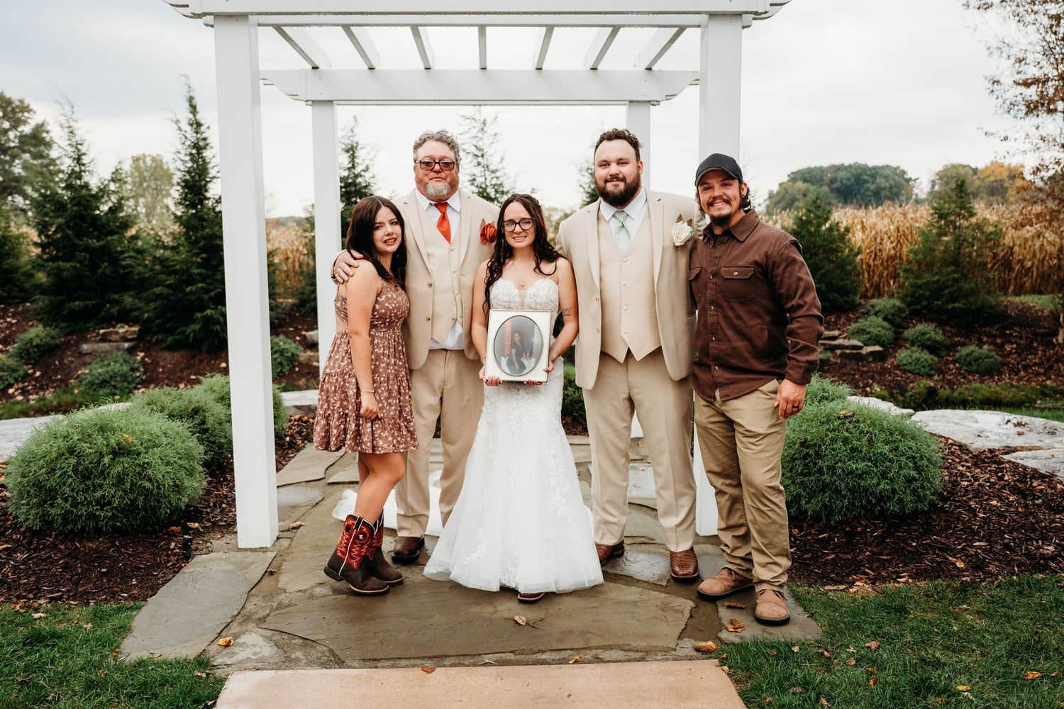 Wedding party of five posing under white pergola in autumn garden, bride in white gown with groomsmen and guests.