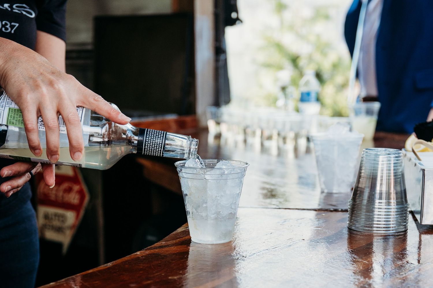 A bartender pours a cocktail from a shaker into a plastic cup at an outdoor wedding bar setup.