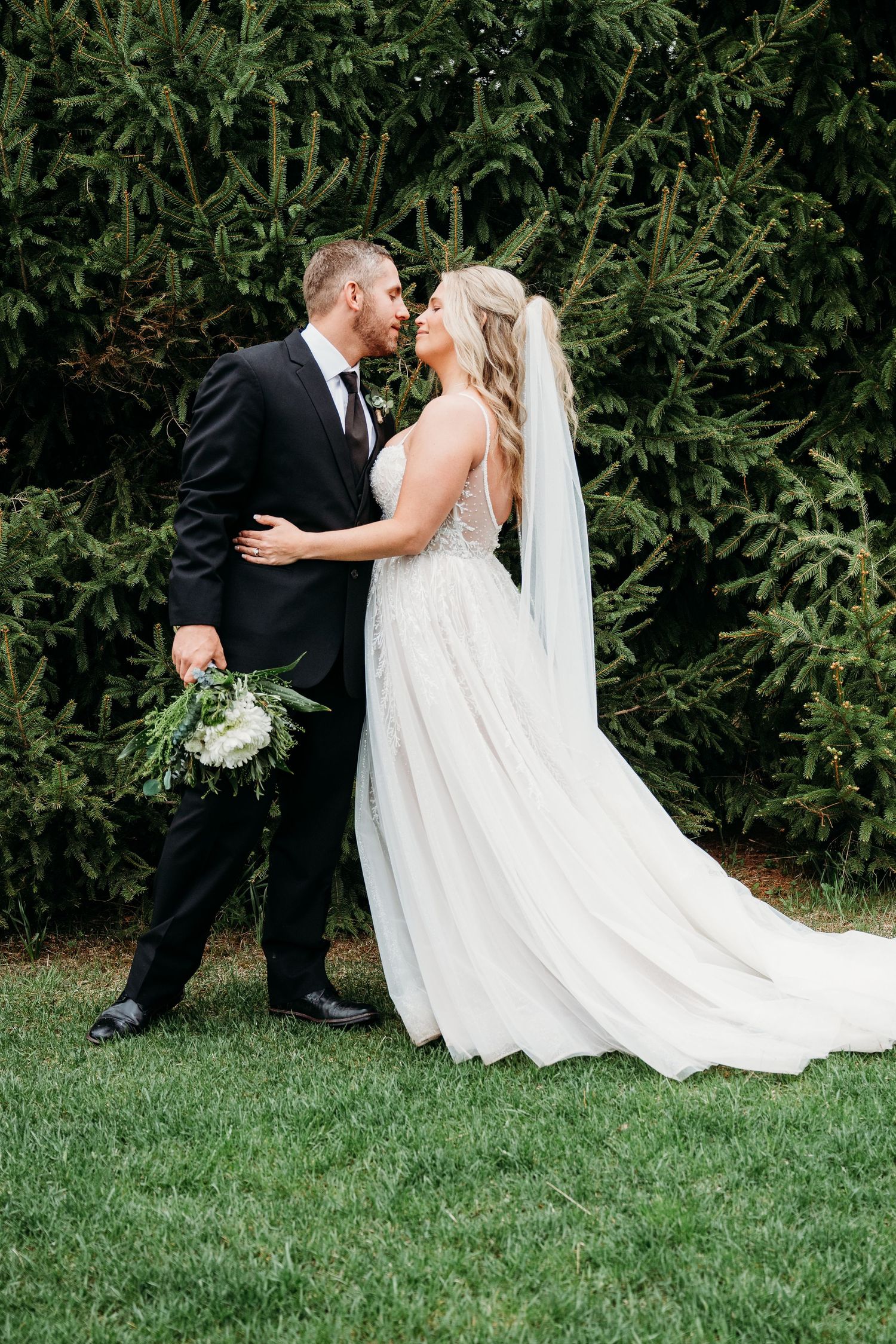 Bride and groom share a romantic kiss in a lush garden, bride wearing a flowing white gown with veil and bouquet.