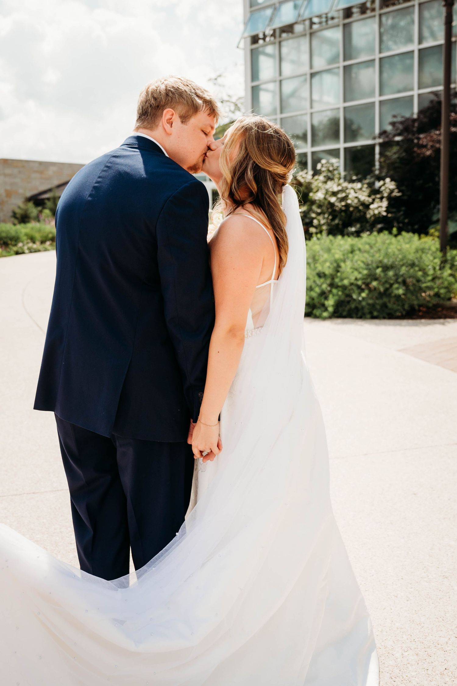 A bride and groom share a kiss outdoors, holding hands, with the bride's white gown flowing in the breeze.