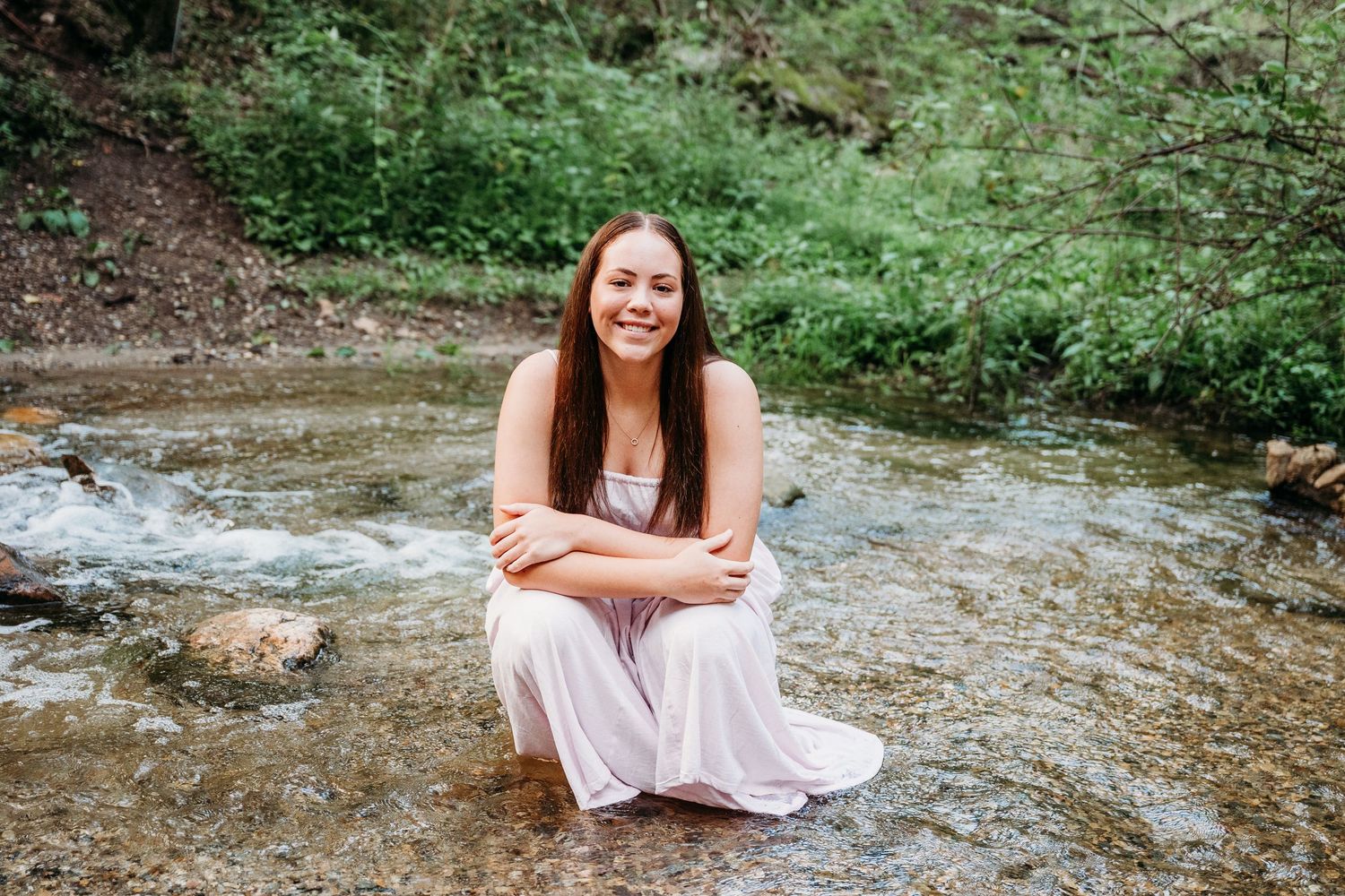 Someone sits by a rocky stream in a flowing pink dress creating ethereal summer portraits amid nature.