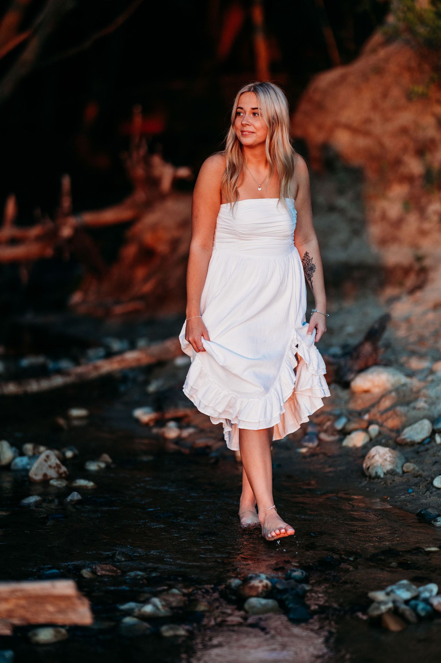 A woman in a flowing white dress stands in a rocky stream during golden hour.