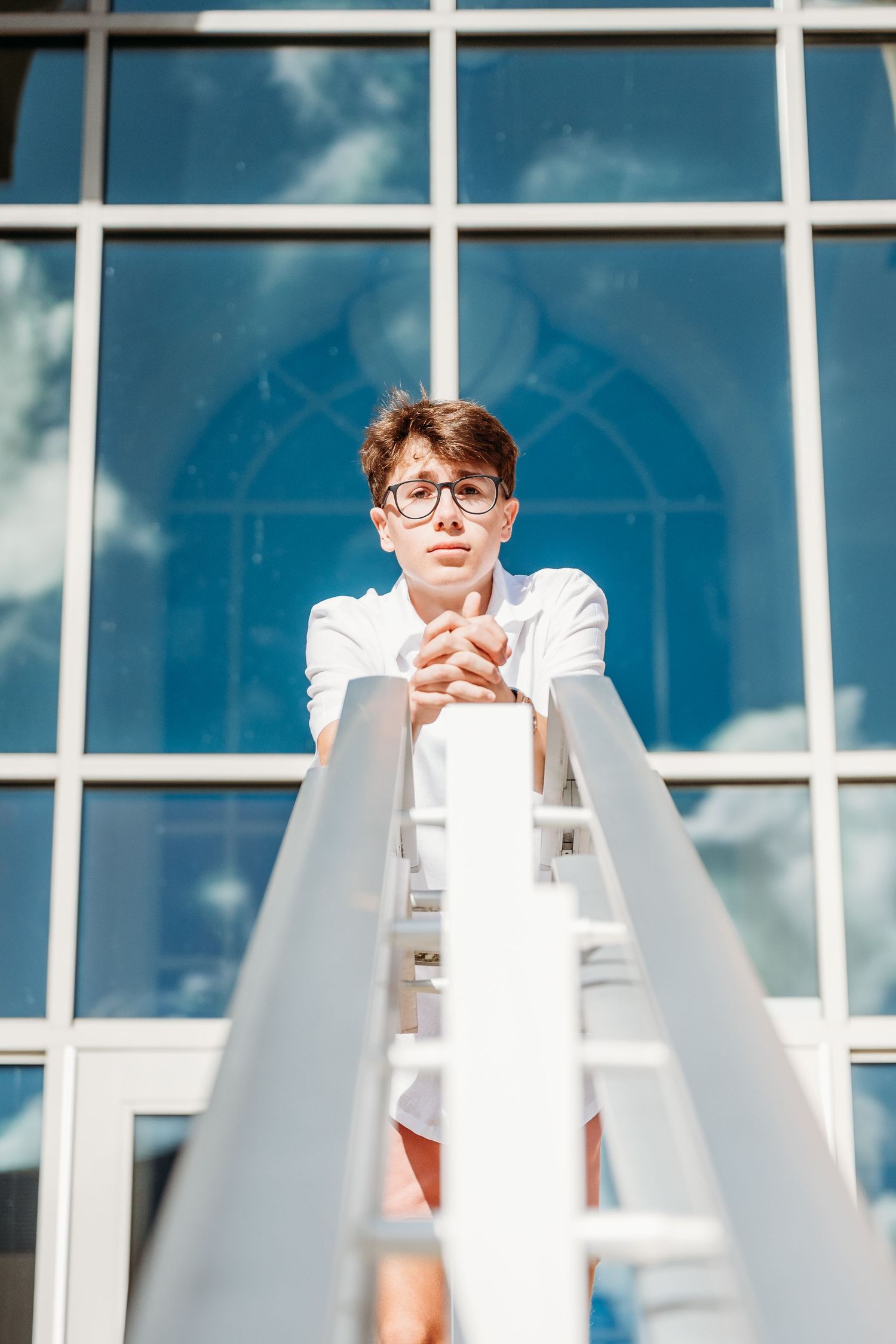 Person wearing white clothing stands between geometric shapes against blue-tinted glass windows and sky backdrop.