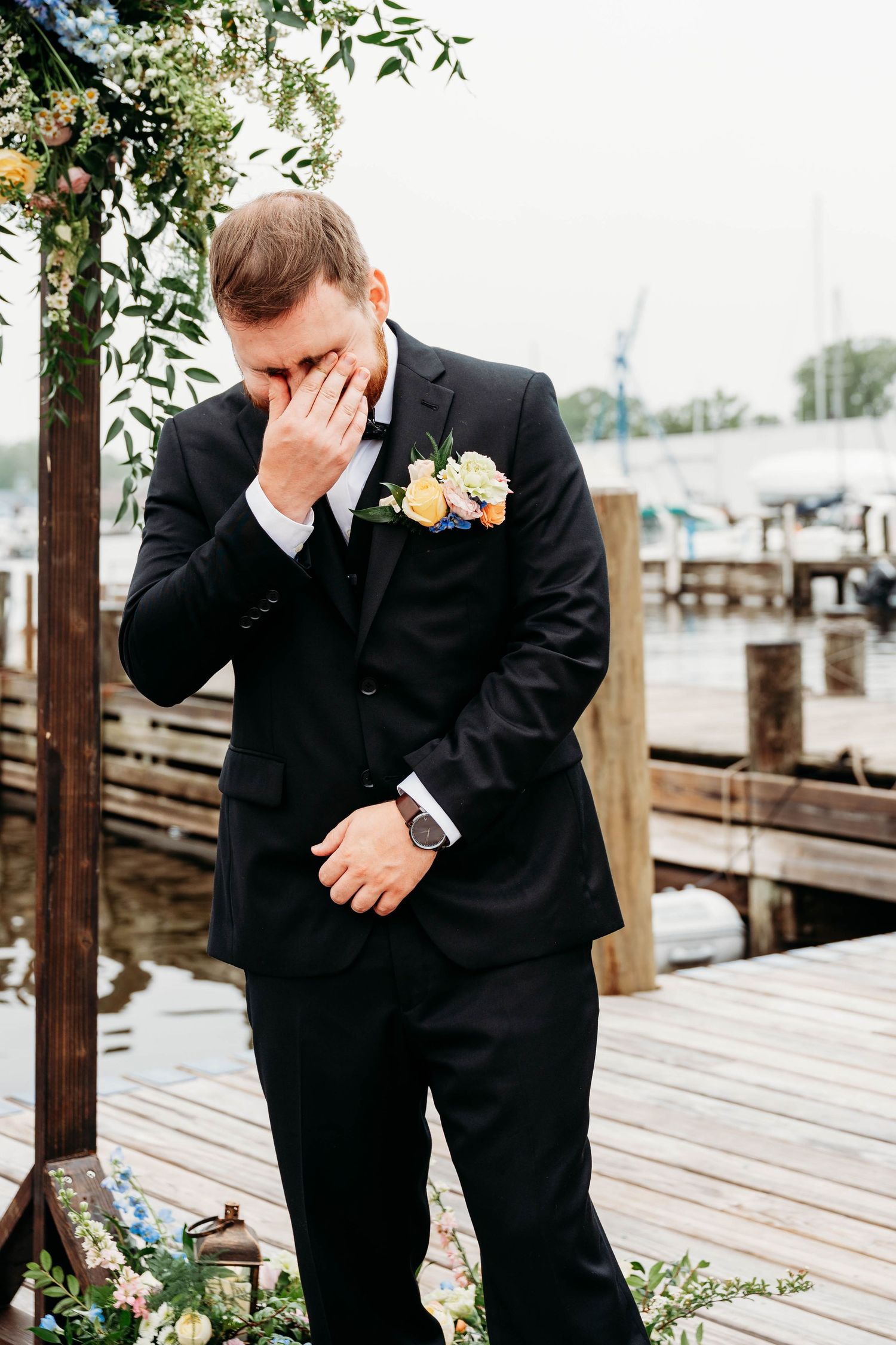 Emotional groom in black tuxedo wiping tears at waterfront wedding ceremony near marina dock.