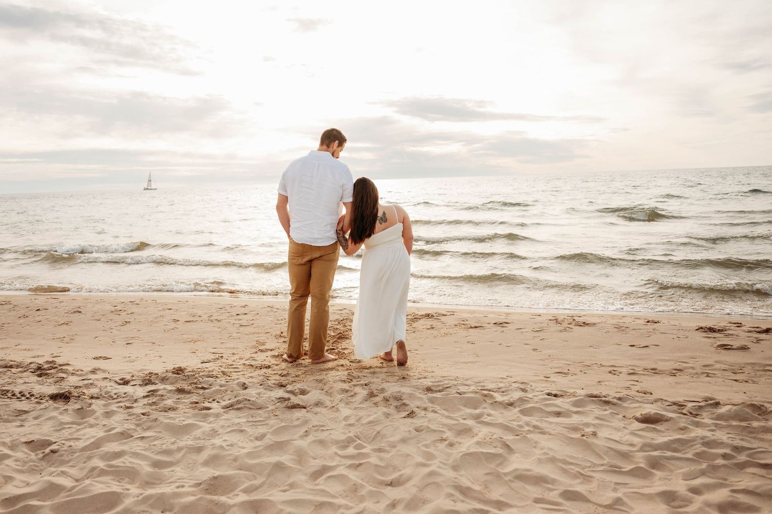 Couple holding hands on sandy beach at sunset, facing the ocean waves in coordinating white and tan outfits.