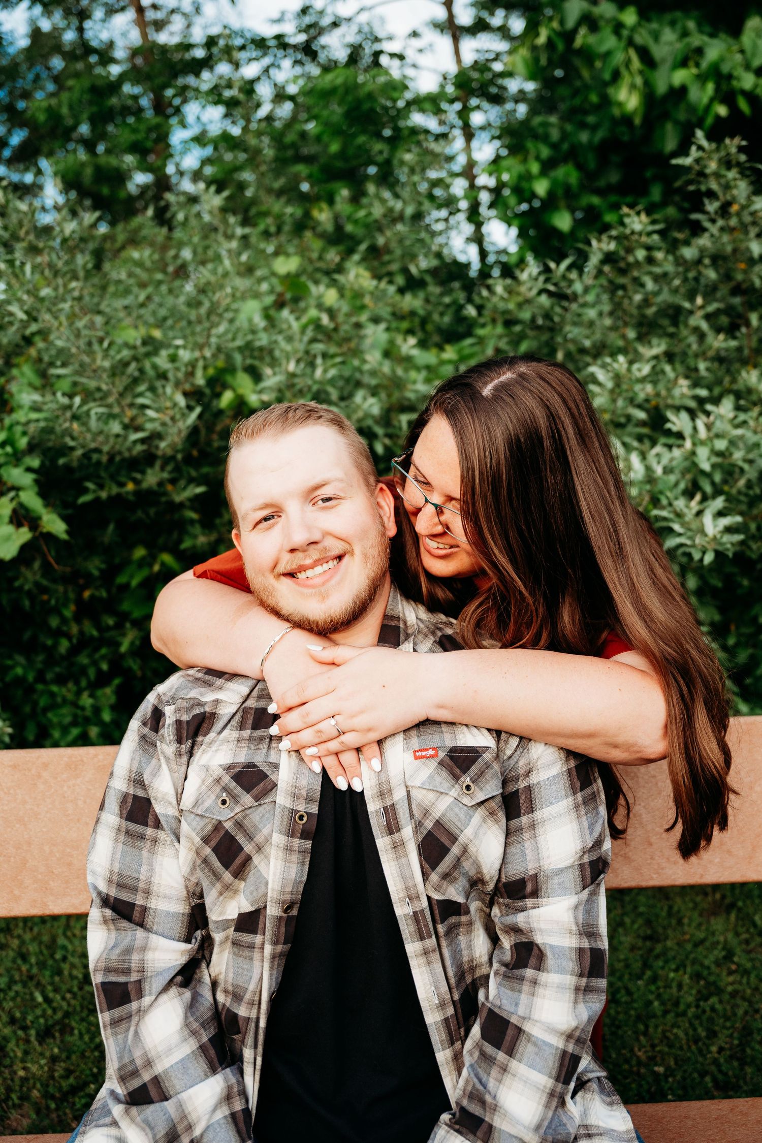 A smiling man in a plaid shirt sits on a bench while a brunette woman hugs him from behind outdoors.