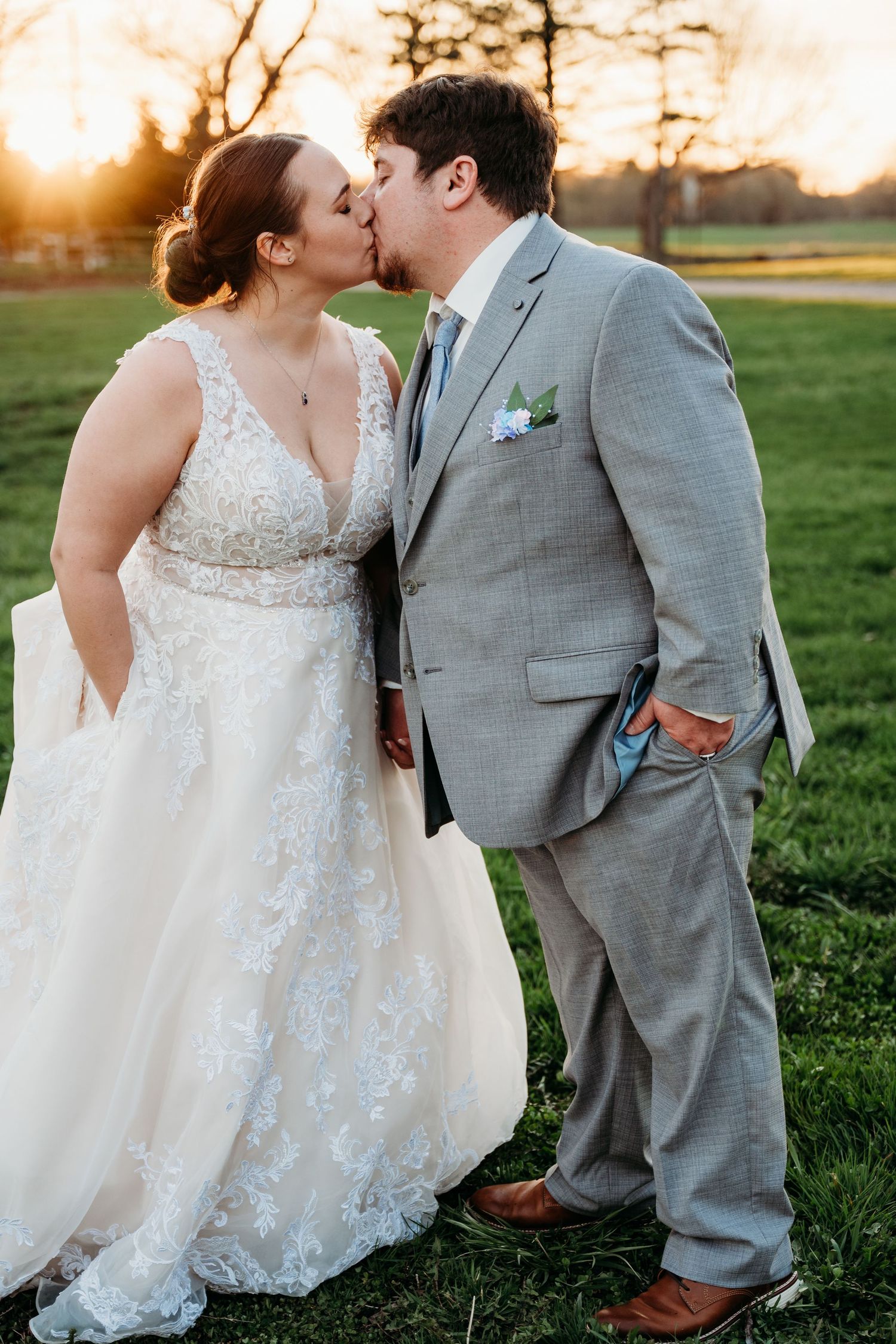 A romantic wedding couple shares tender moments in a grassy field at sunset, with the bride in lace and groom in gray suit.