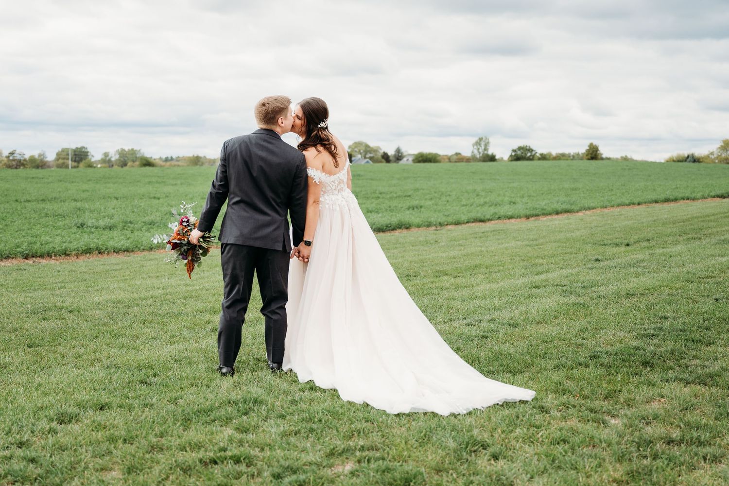 A bride and groom share a kiss while holding hands in a lush green field on their wedding day.