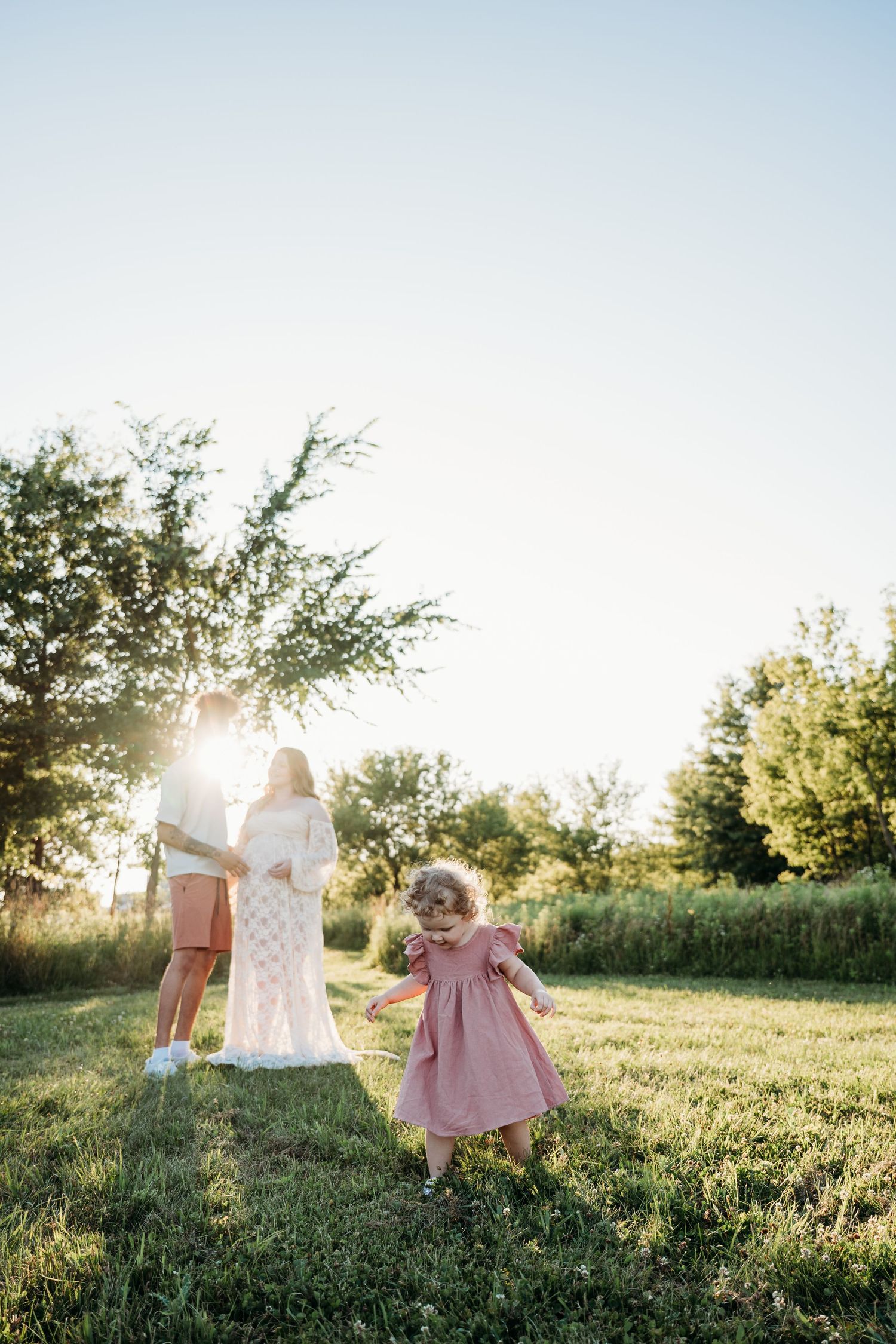 A family moment captured in golden sunlight with a small child in a pink dress running joyfully on grass.