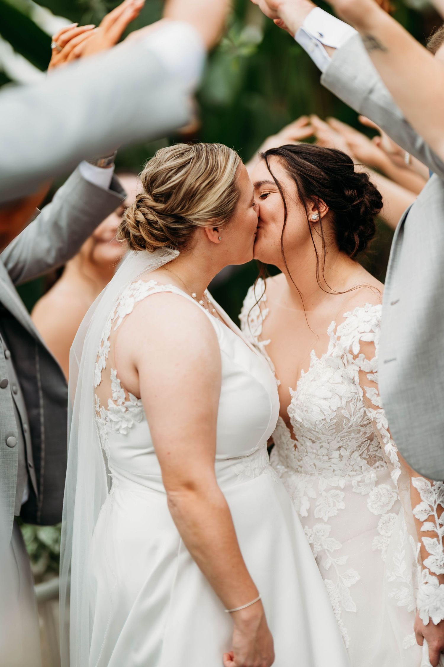 Two brides in white wedding dresses share a romantic moment under raised arms of celebrating guests.