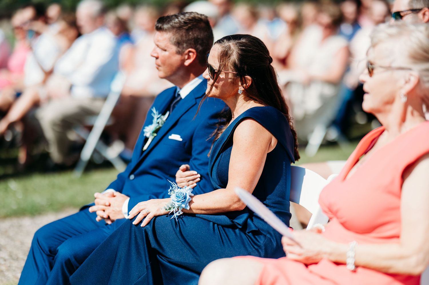 Wedding guests in blue and coral attire sit together during an outdoor ceremony.