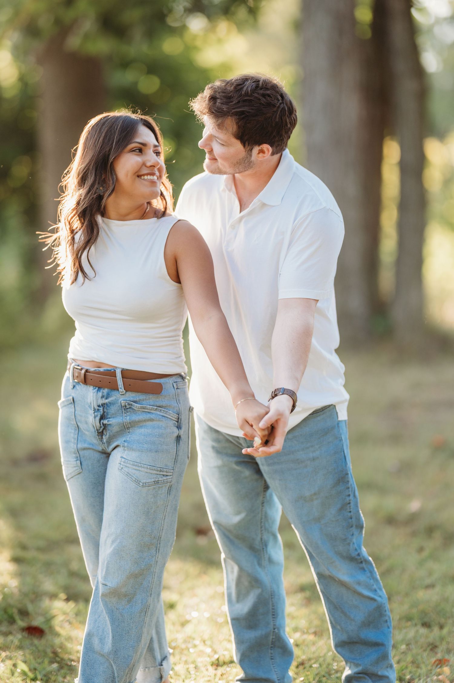 Couple in white tops and blue jeans posing playfully together in a sunlit forest setting.