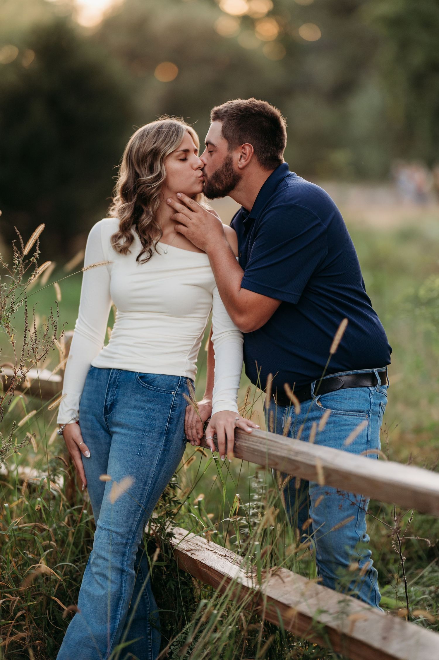 Romantic couple sharing tender moments along a wooden fence in a sunlit meadow with tall grass and wildflowers.
