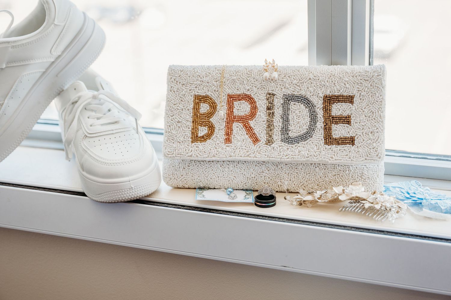 White sequined clutch purse with 'BRIDE' text in gold sits on windowsill next to white sneakers.