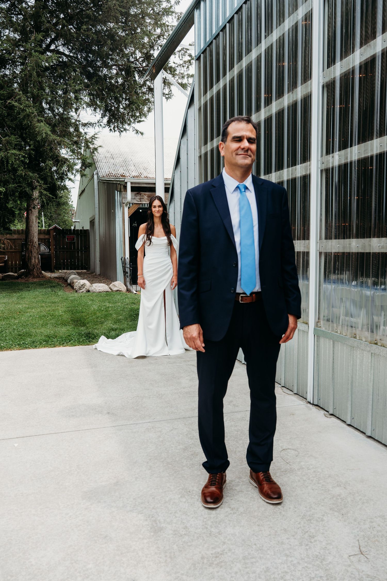 A wedding scene with two people, one in a navy suit and blue tie and another in an elegant white dress by a modern building.