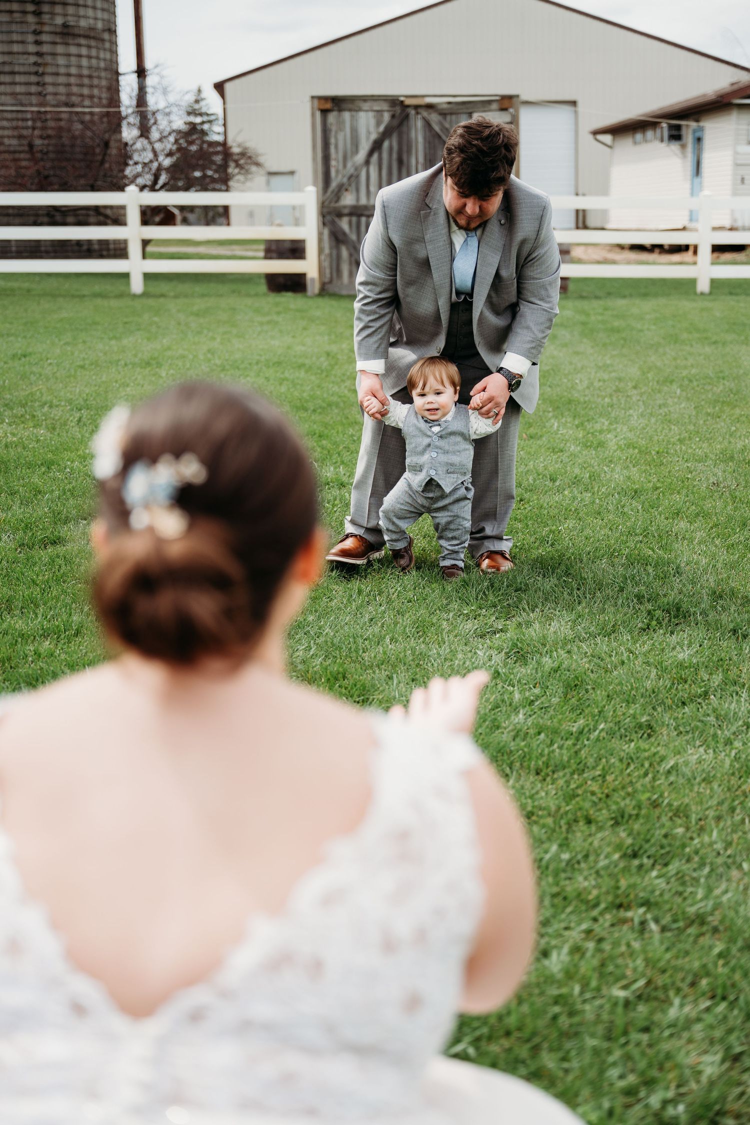 Adult helping small child take first steps on green lawn while someone in wedding dress watches from behind.