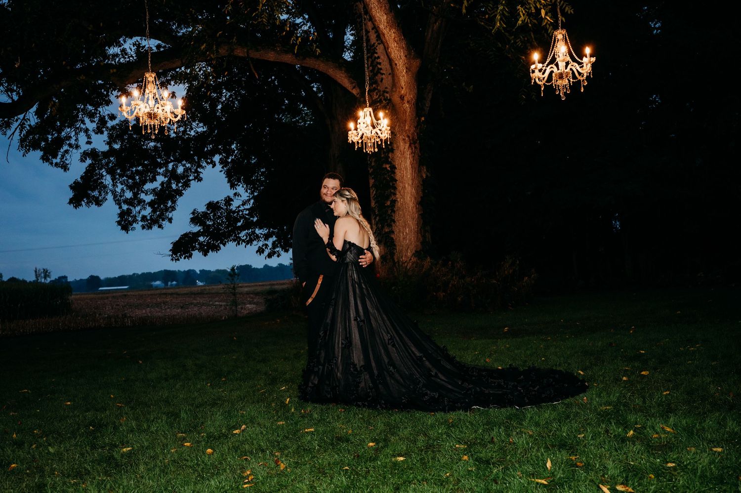 Silhouetted romantic couple stands beneath trees at dusk as warm light streams through branches during sunset photoshoot.