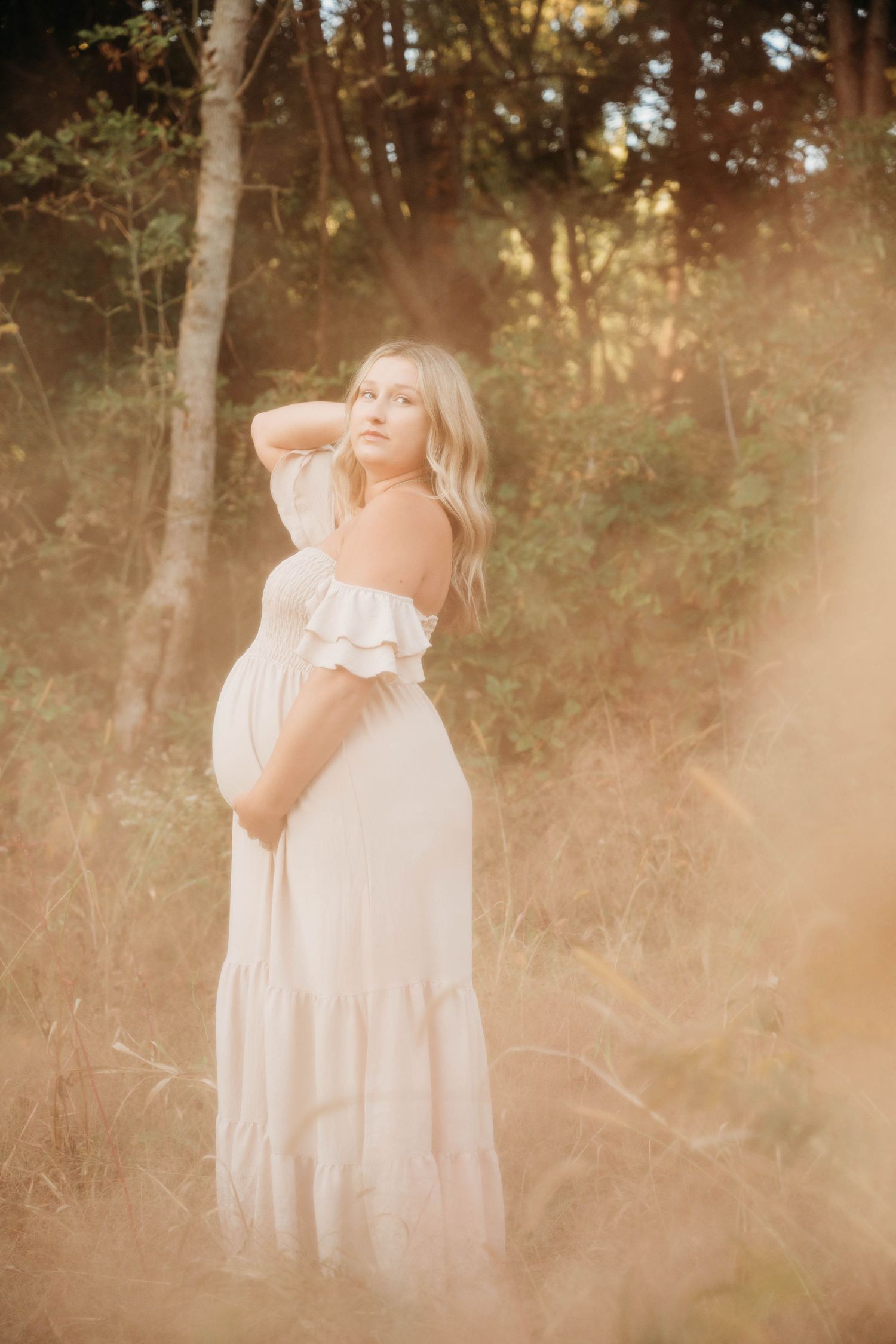 Ethereal maternity photo in a dreamy field at golden hour with soft light filtering through trees.