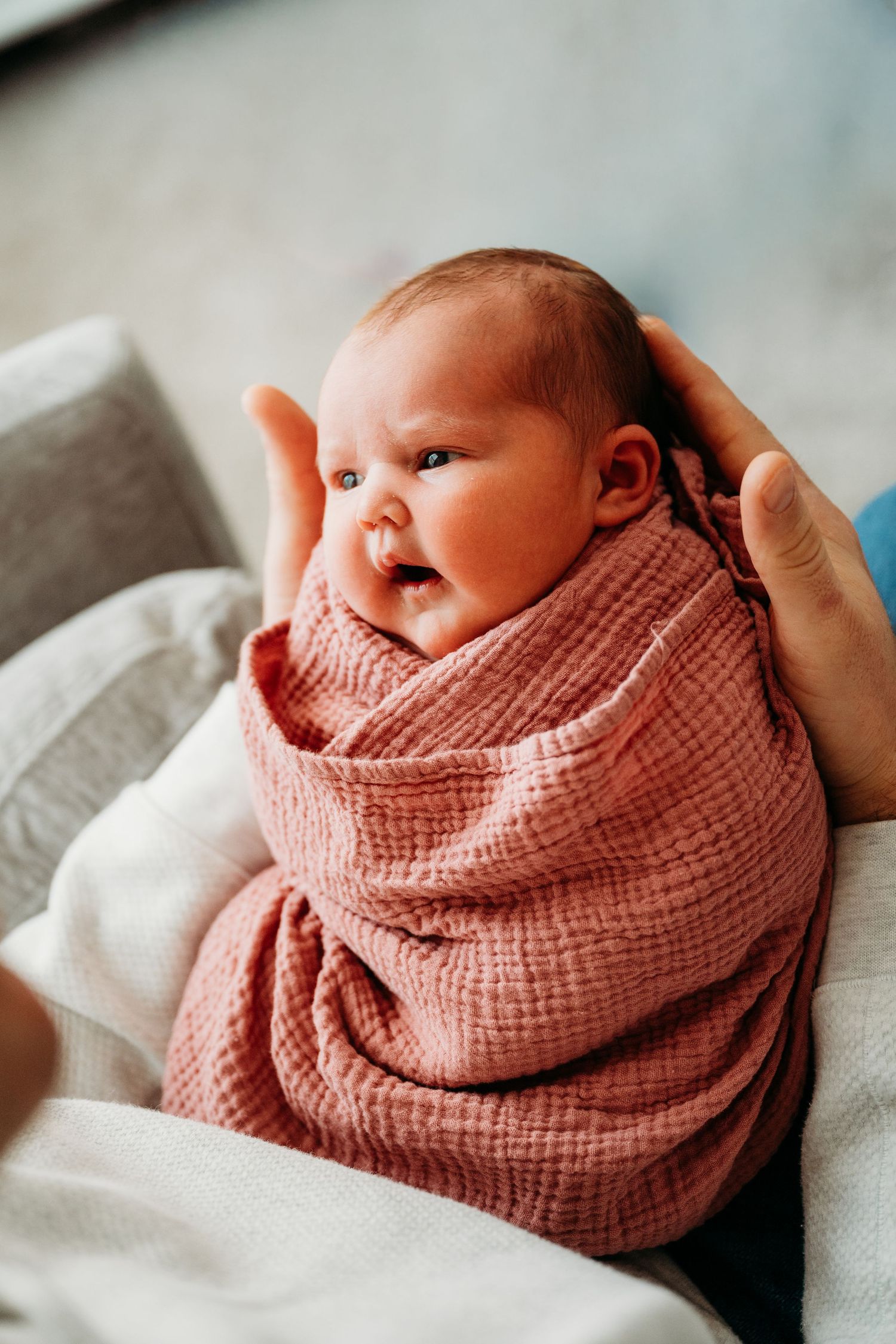 A newborn baby wrapped in a soft pink muslin blanket rests on white bedding while being gently cradled.