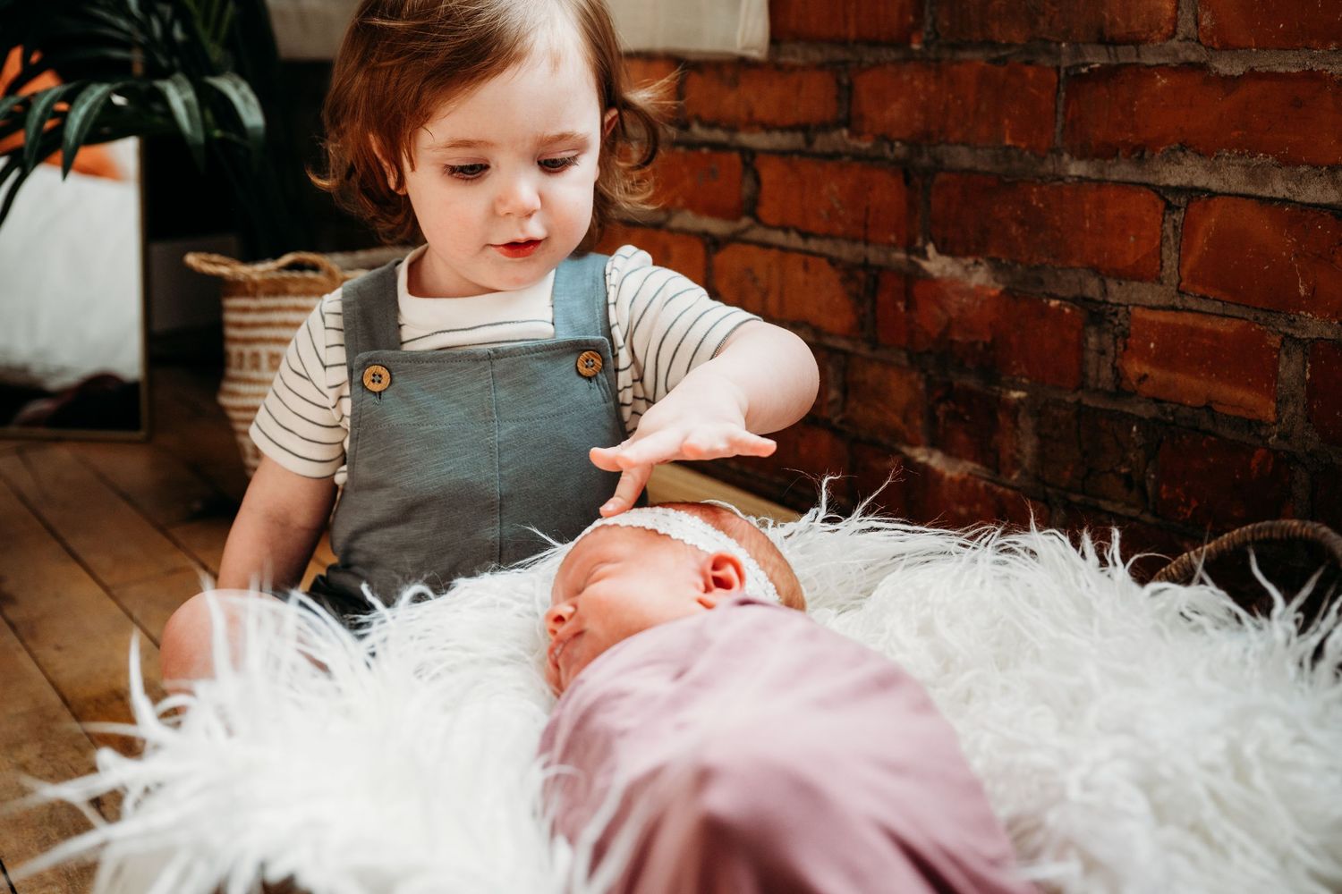 Baby lying on soft white fur blanket while sibling in gray overalls looks on against brick wall background.