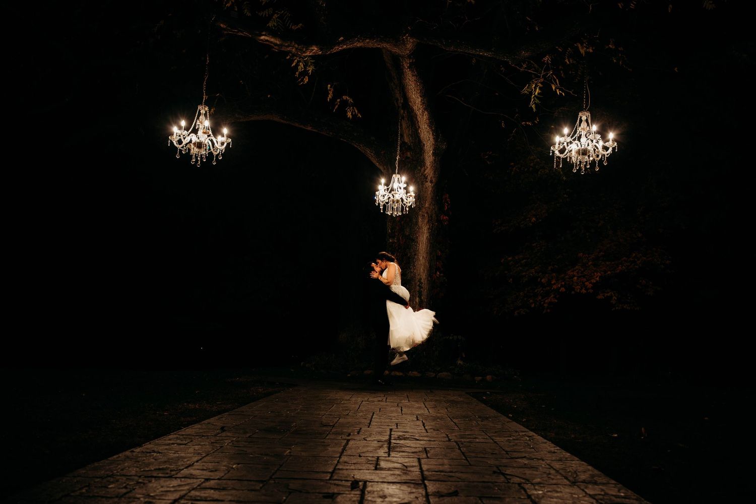 A dark cobblestone path lit by ornate chandeliers leads through an atmospheric tunnel at night.