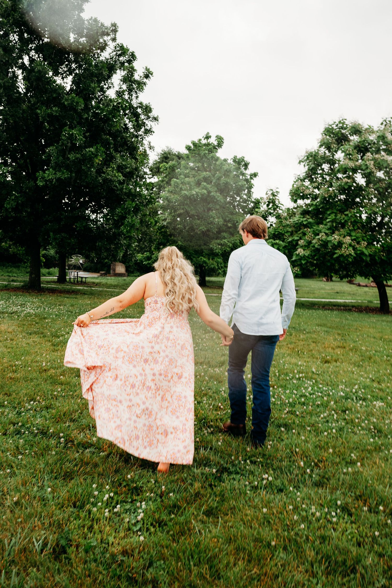 Couple walking hand-in-hand through lush green park, woman in floral pink maxi dress, man in light blue shirt.