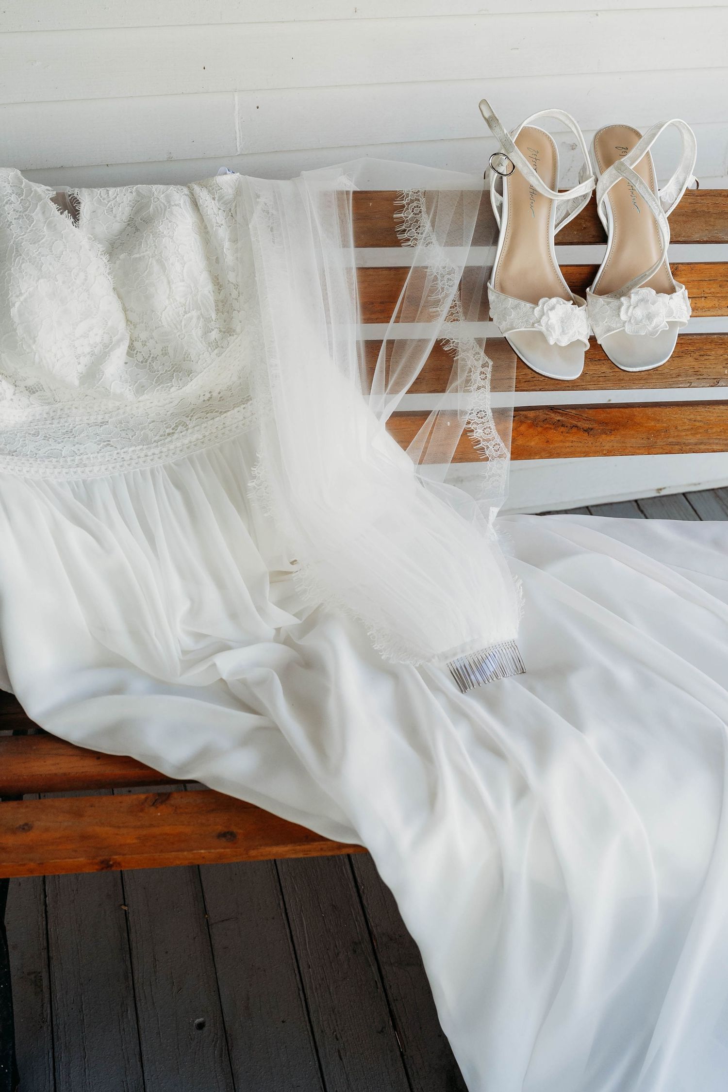 White lace wedding dress with veil and floral heeled sandals displayed on a wooden bench.