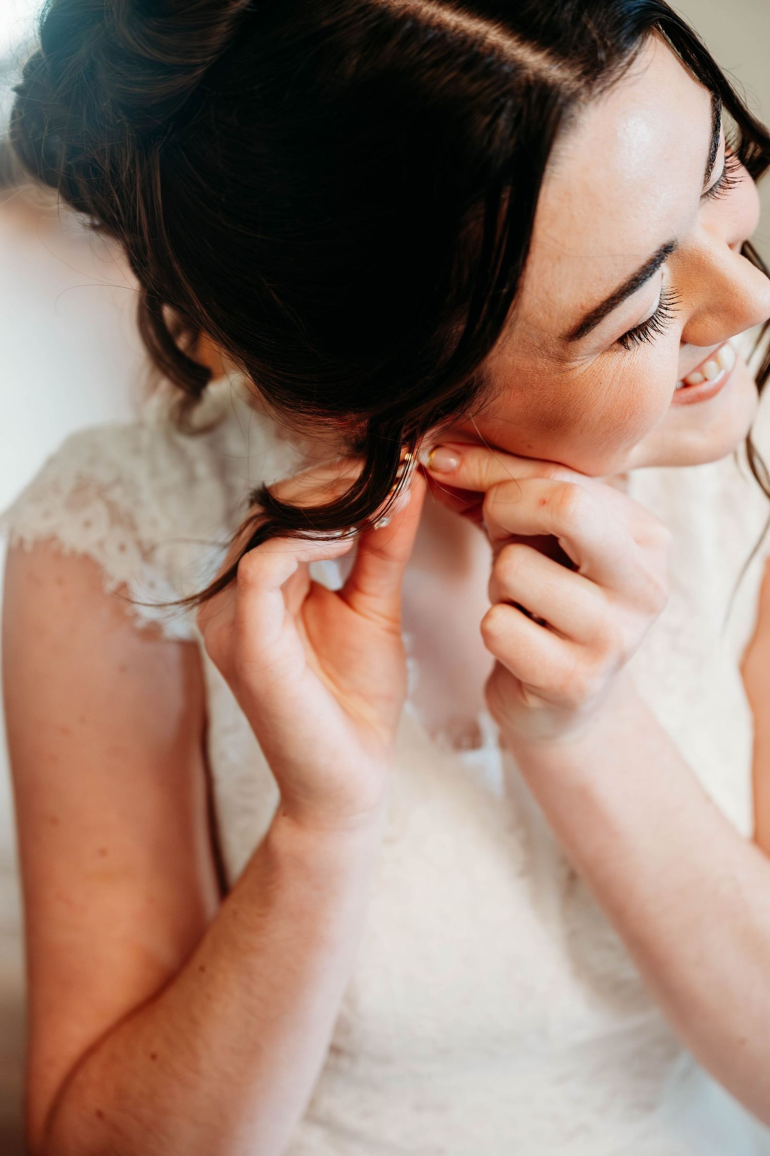 Bride in white lace dress smiling while putting on earrings on her wedding day.