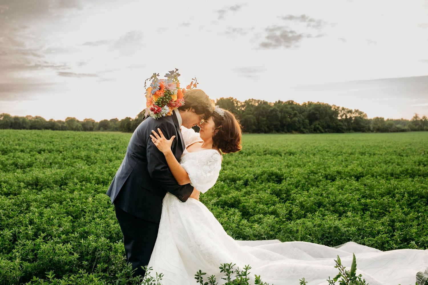 Bride and groom sharing a romantic kiss in a lush green field at sunset, bride holding a colorful bouquet.