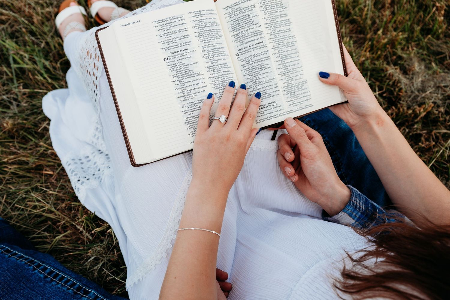 Woman lying in grass reading an open Bible, wearing white top and jeans, with blue nail polish and a ring.